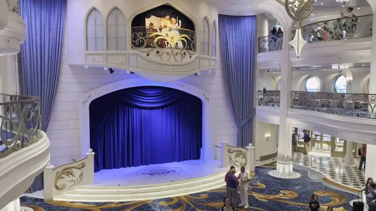 A grand, elegant atrium features a small stage with rich blue curtains and ornate railings. Above, a balcony with a decorative facade overlooks the area. People are scattered across the multi-level space, which includes plush carpeting and intricate light fixtures.