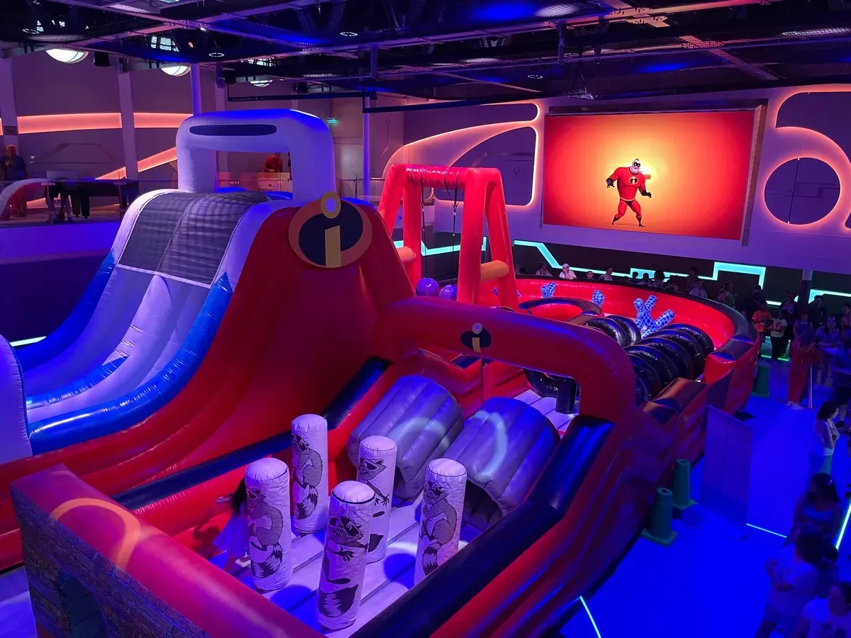 A vibrant indoor inflatable obstacle course themed around "The Incredibles" features a large slide and various challenges. The setup includes red and blue inflatables with the iconic "i" logo, surrounded by colorful lighting. In the background, a large image of a superhero character adds to the energetic atmosphere.