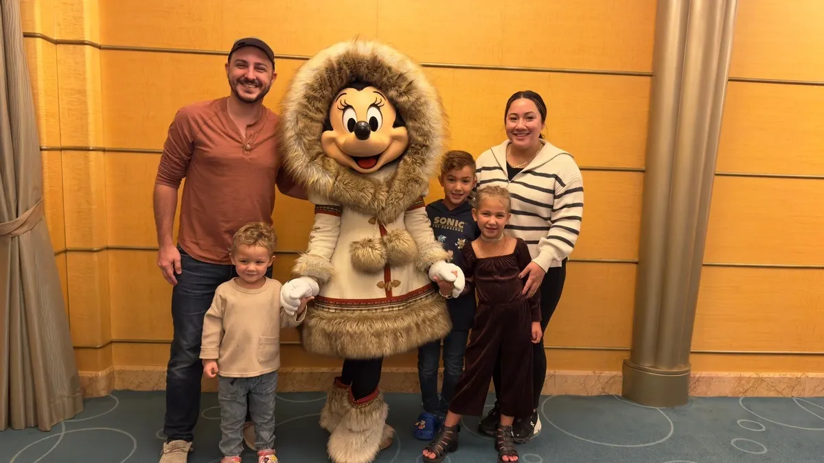 A family of five poses with Minnie Mouse, who is dressed in a cozy fur-trimmed parka. The family stands in a warmly lit room with a beige wall and blue patterned carpet. The children are smiling, and the atmosphere is joyful and friendly.