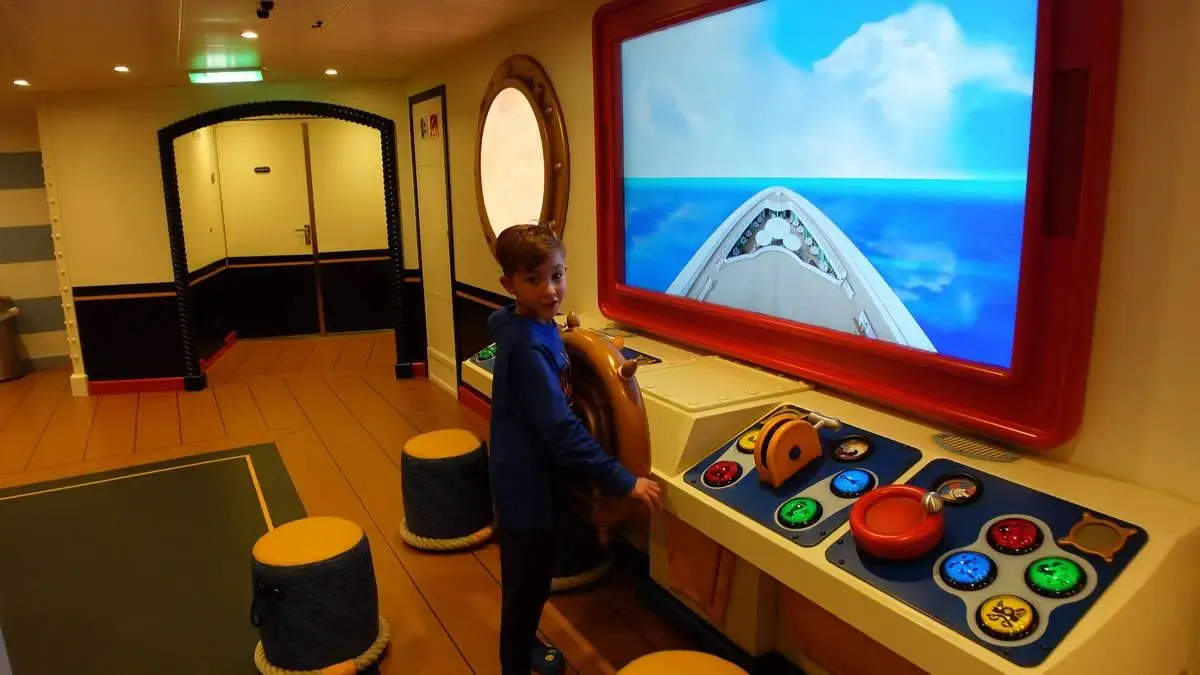 A child in a blue shirt is playing with a ship simulator, steering a wooden wheel in front of a large screen displaying an ocean scene. The room has a nautical theme, with porthole windows and colorful control panels featuring various buttons and levers.
