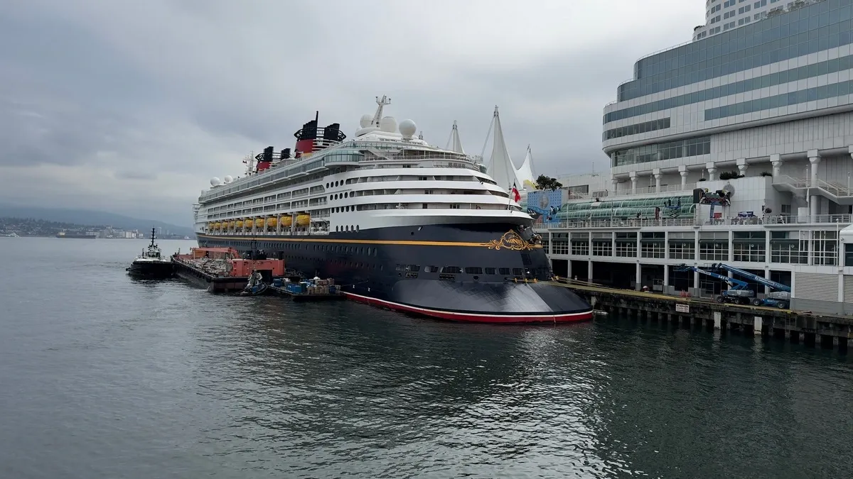A large cruise ship is docked at a bustling port, with a tugboat nearby assisting. The ship features a dark hull with a decorative emblem and multiple decks with yellow lifeboats. The port building is modern and expansive, set against a cloudy sky.