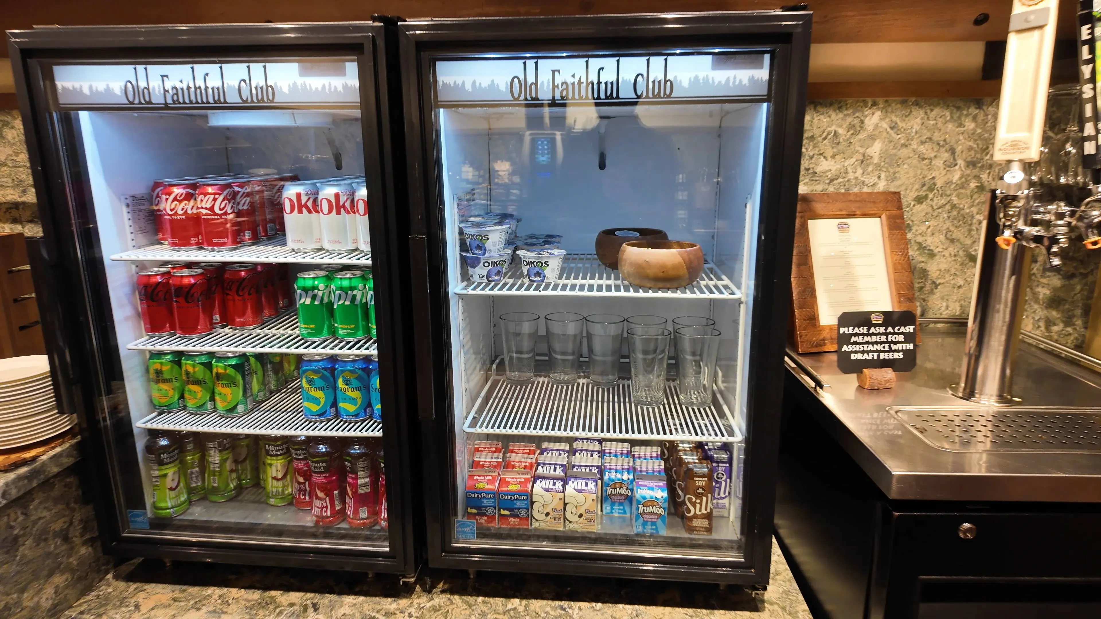 Two glass refrigerators display an assortment of beverages, such as Coca-Cola, Diet Coke, Sprite, and various juices. The right fridge contains yogurt cups, milk cartons, and a few empty glasses. A sign next to the fridges offers assistance with draft beers.