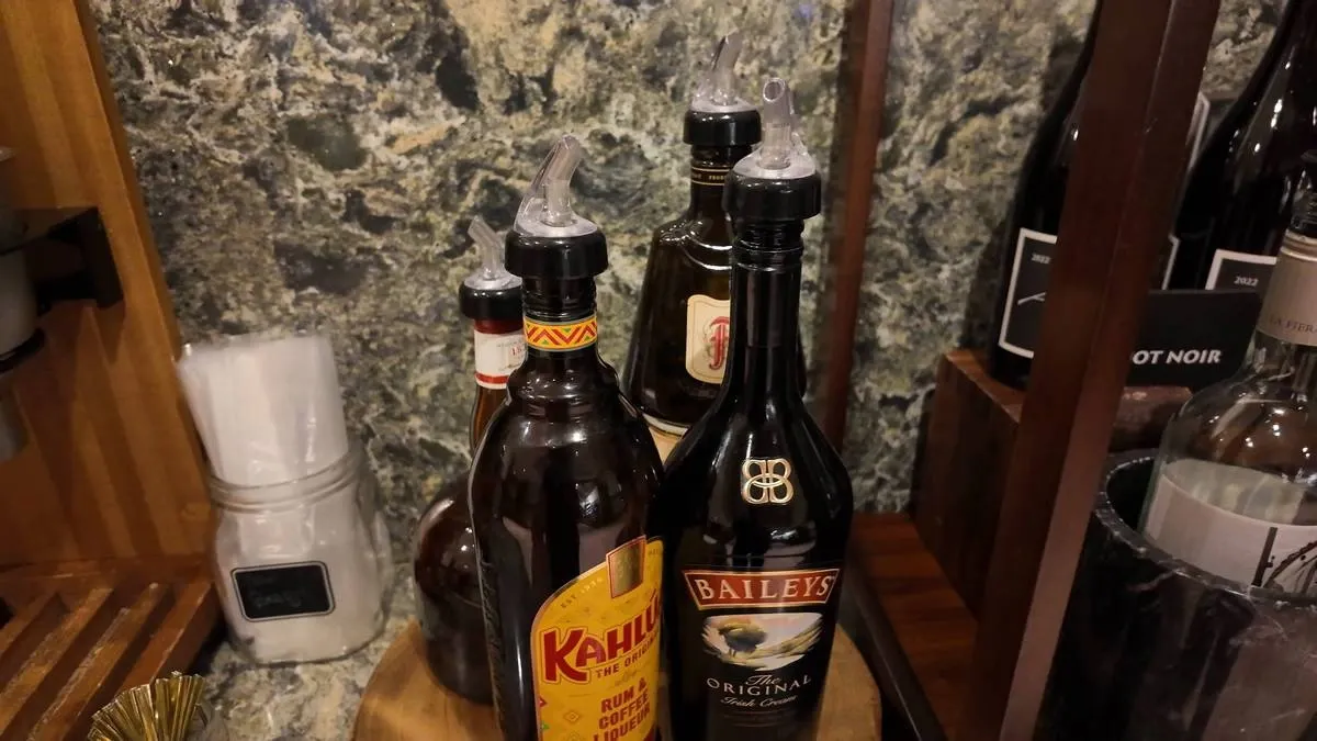 A selection of liquor bottles, including Kahlúa and Baileys, are arranged on a wooden tray against a textured stone wall. Nearby, a jar with white napkins sits on the counter, adding to the bar setup.