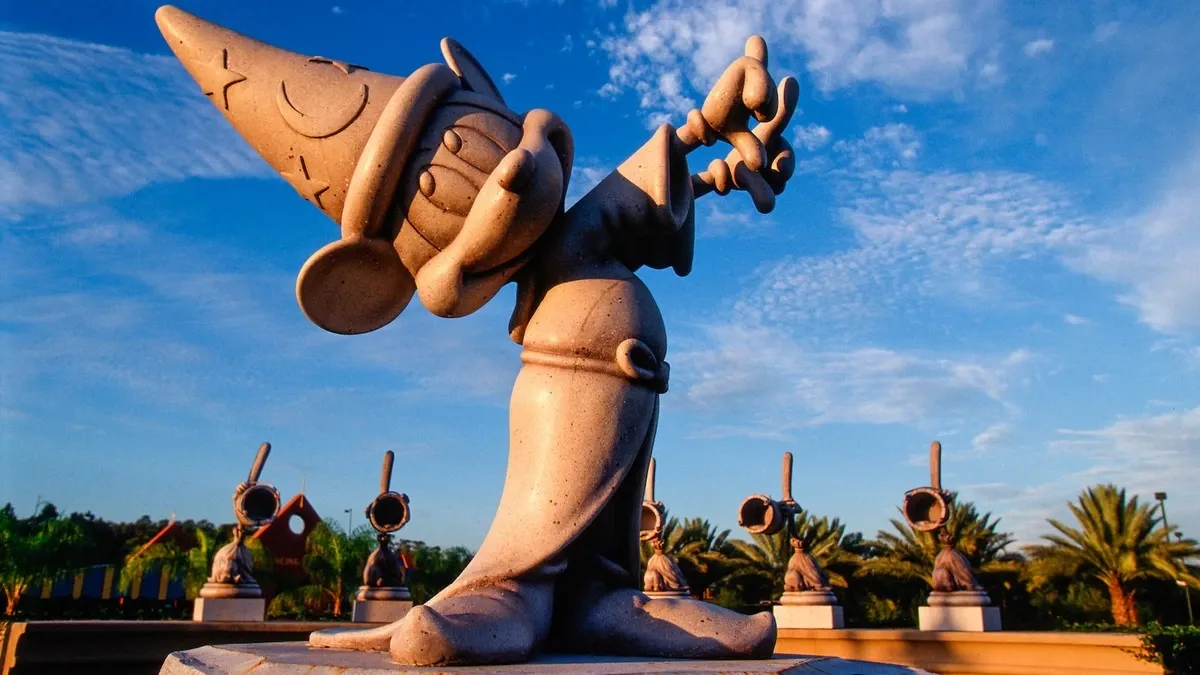 A whimsical statue of a cartoon character wearing a wizard hat and robe stands prominently under a bright blue sky, surrounded by palm trees and smaller similar statues in the background.