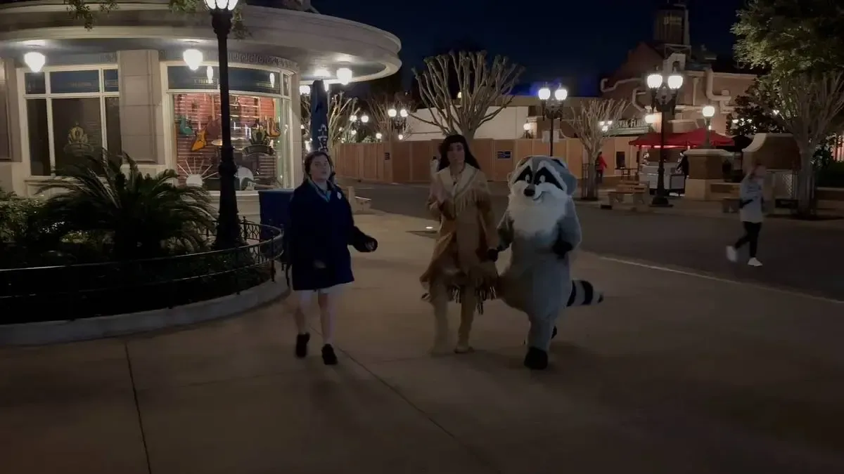 A person dressed as Pocahontas walks hand in hand with a person in a raccoon costume through a nighttime theme park. A woman in a blue jacket walks beside them, and a round building with bright windows is in the background.