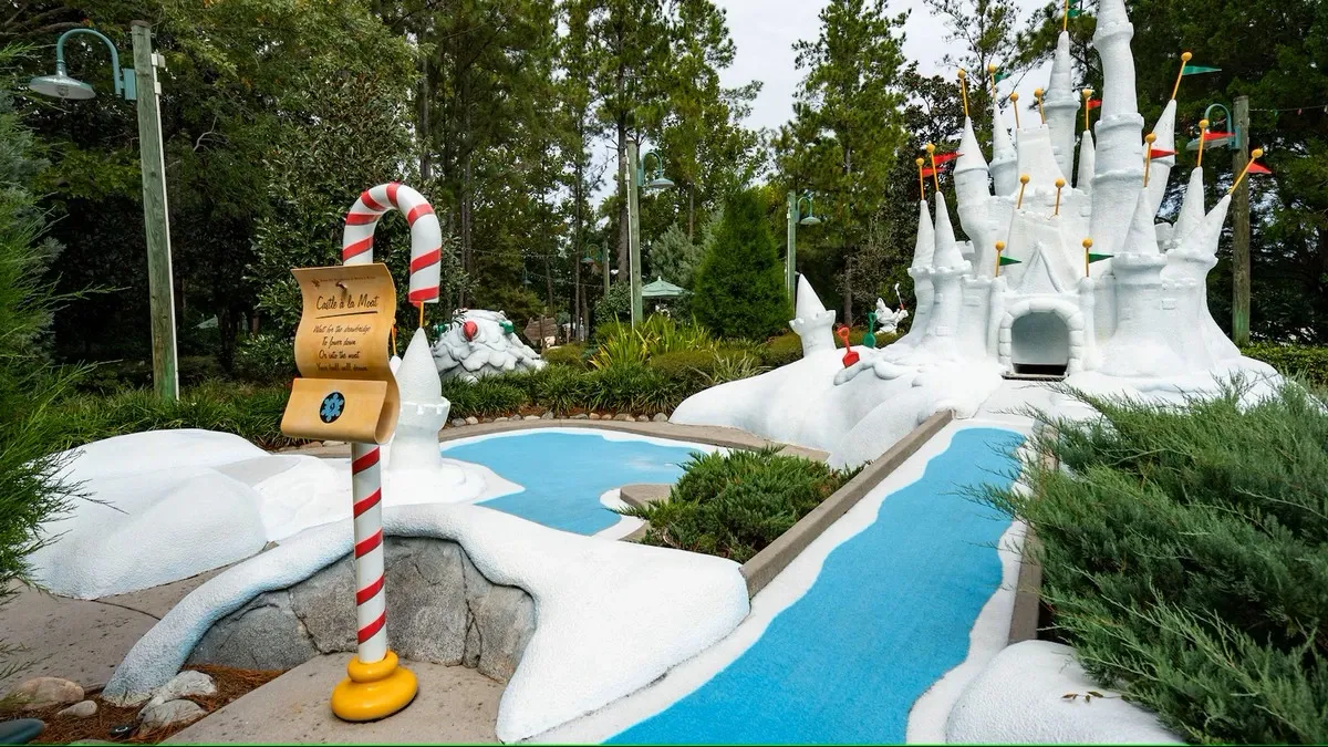 A mini-golf course features a candy cane signpost with a playful message, set against a backdrop of a castle covered in snow adorned with colorful flags.