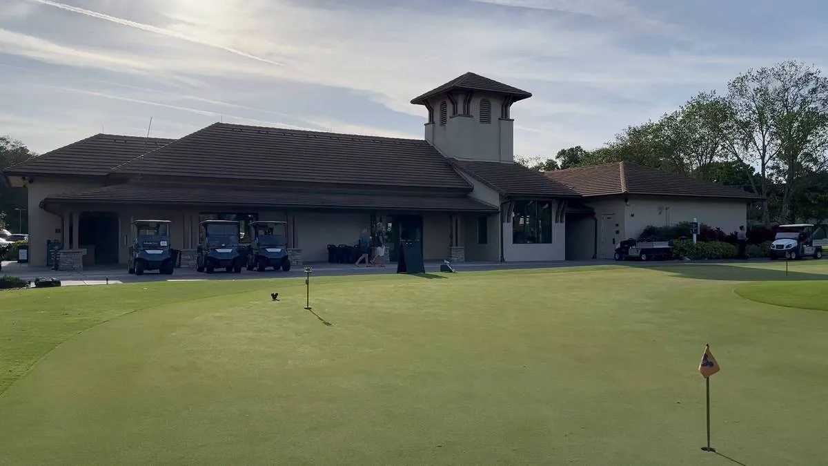 A golf clubhouse with a brown tiled roof stands in front of a putting green, with several golf carts parked nearby and trees in the background under a partly cloudy sky.