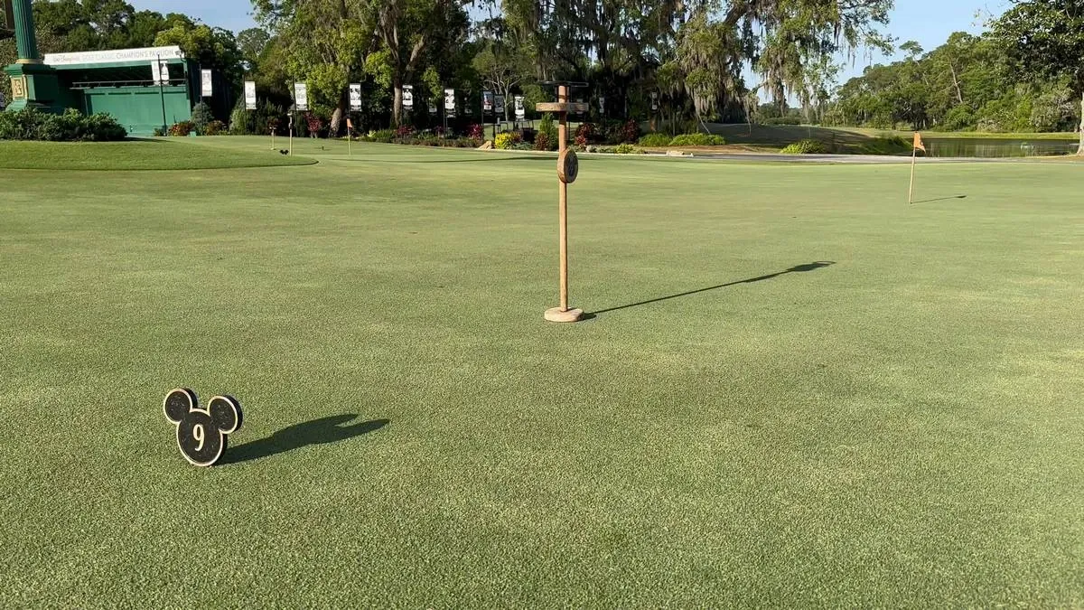 A golf course green features a Mickey Mouse-shaped marker with the number 9, surrounded by lush greenery and trees in the background. A flagstick is visible, indicating the hole's location.