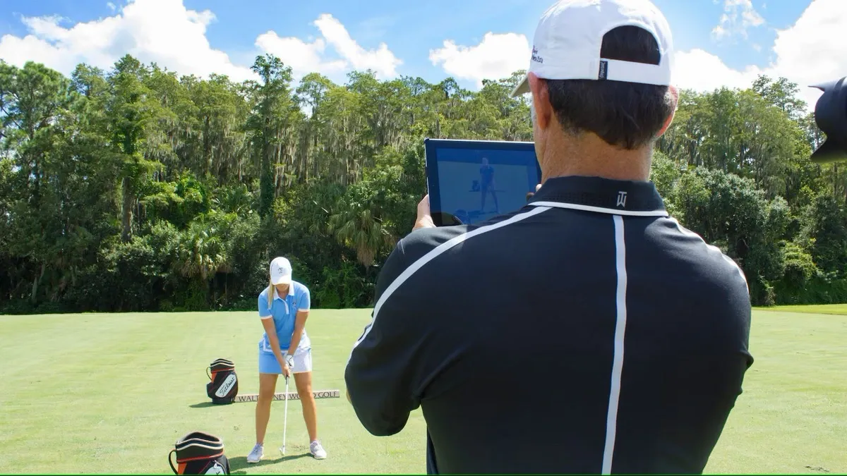 A golf coach uses a tablet to record a player in a light blue outfit preparing to swing on a lush green golf course, surrounded by tall trees under a bright blue sky.