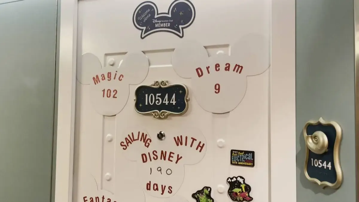 A decorated door features Mickey Mouse-shaped magnets with phrases like "Magic 102," "Dream 9," and "Sailing with Disney 190 days." The room number is prominently displayed in an ornate plaque. There are many small stickers and a colorful train image.