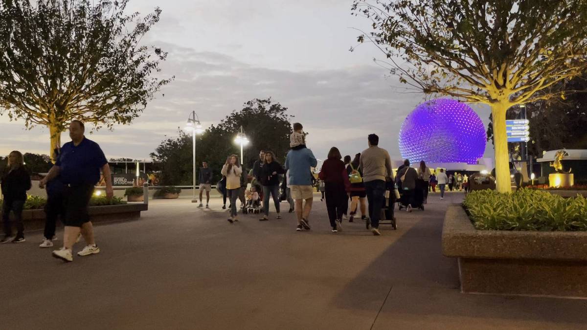 People stroll along a pathway at dusk, with trees lining the sides and a large, and Epcot's sphere illuminated in purple in the background.