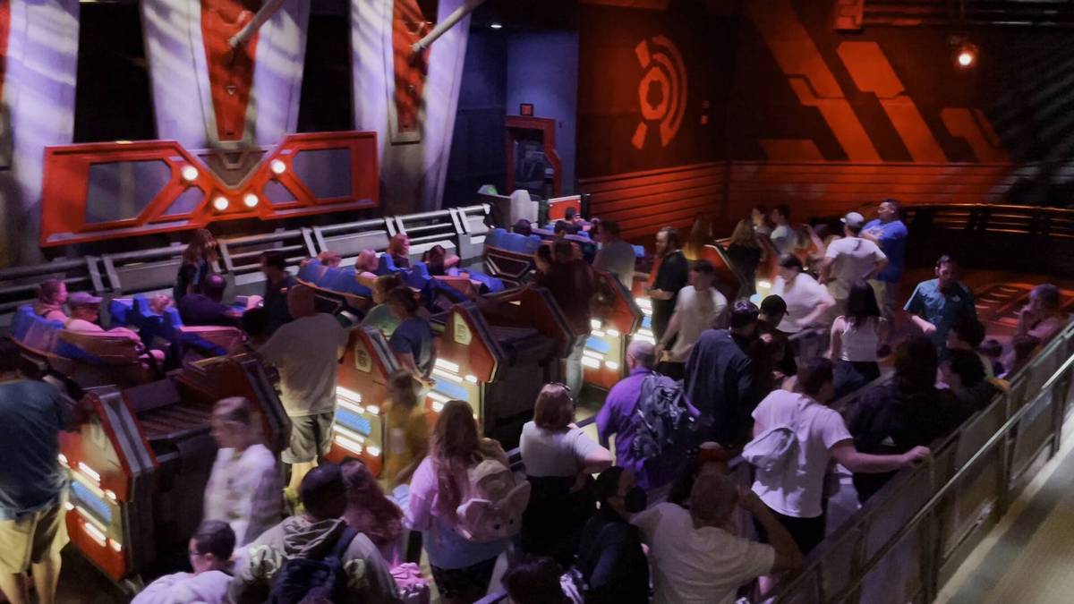 People are boarding a futuristic ride with vibrant red and blue vehicles inside a dimly lit theme park attraction, surrounded by a bustling crowd and colorful lighting effects.