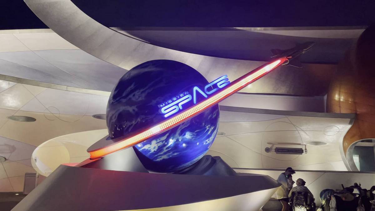 A futuristic sign for "Mission: SPACE" features a blue Earth globe with a sleek red and white rocket orbiting around it, set against a modern architectural background with curved metallic surfaces.