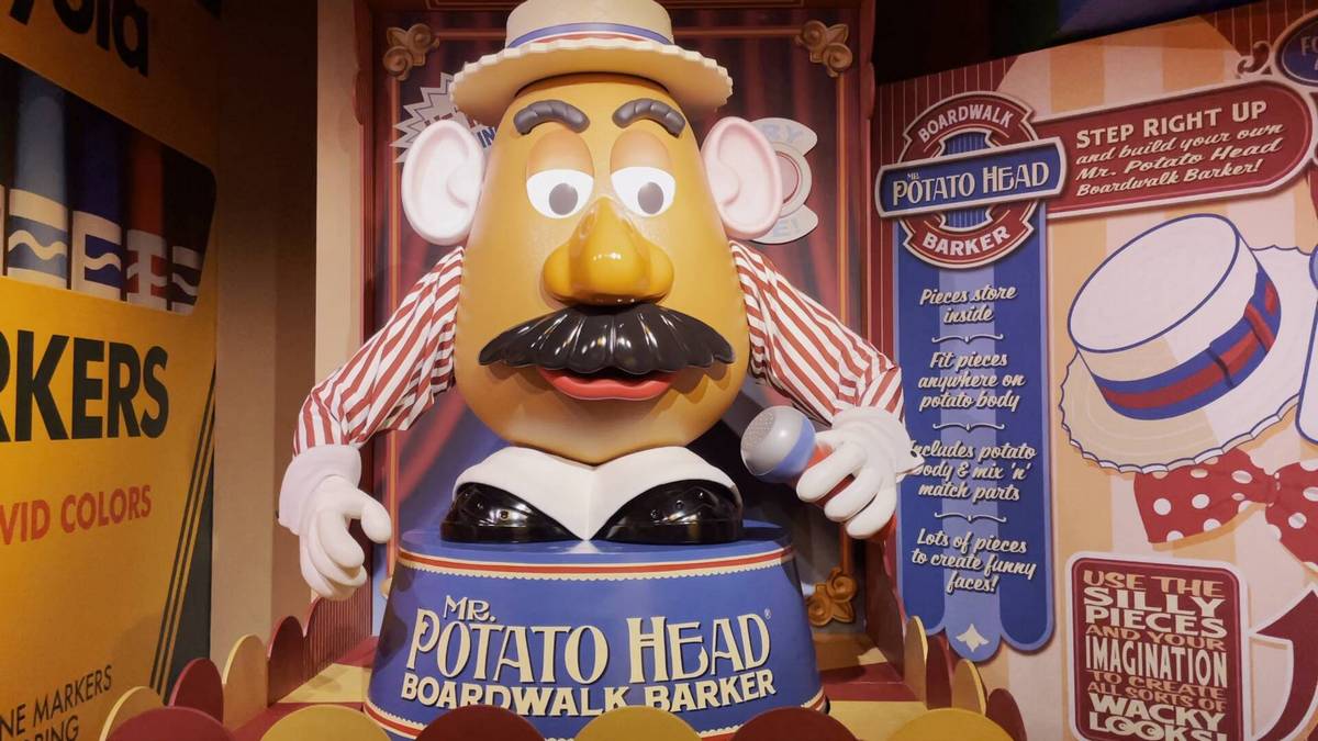 A large Mr. Potato Head figure dressed as a boardwalk barker holds a microphone, wearing a striped shirt and hat. The display includes colorful signage inviting people to build their own Mr. Potato Head with various mix-and-match parts.