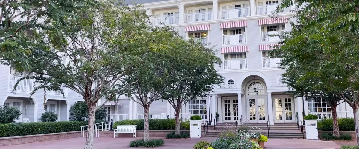A charming hotel facade with white siding and pink-striped awnings, surrounded by green trees and shrubs, featuring a welcoming entrance with large glass doors and a few white benches in front.