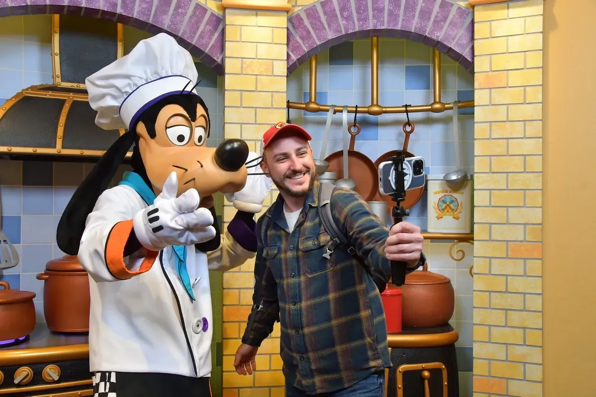 A person takes a selfie with a character dressed as a chef, wearing a white hat and coat, in a colorful kitchen setting with hanging pots and pans.