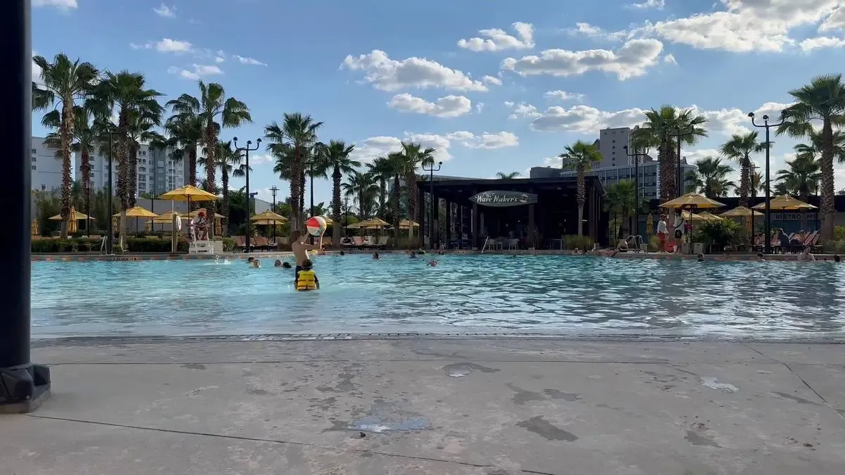 A sunny outdoor pool scene with people swimming and playing under a clear blue sky. Palm trees and yellow umbrellas line the poolside, adding a tropical vibe. 