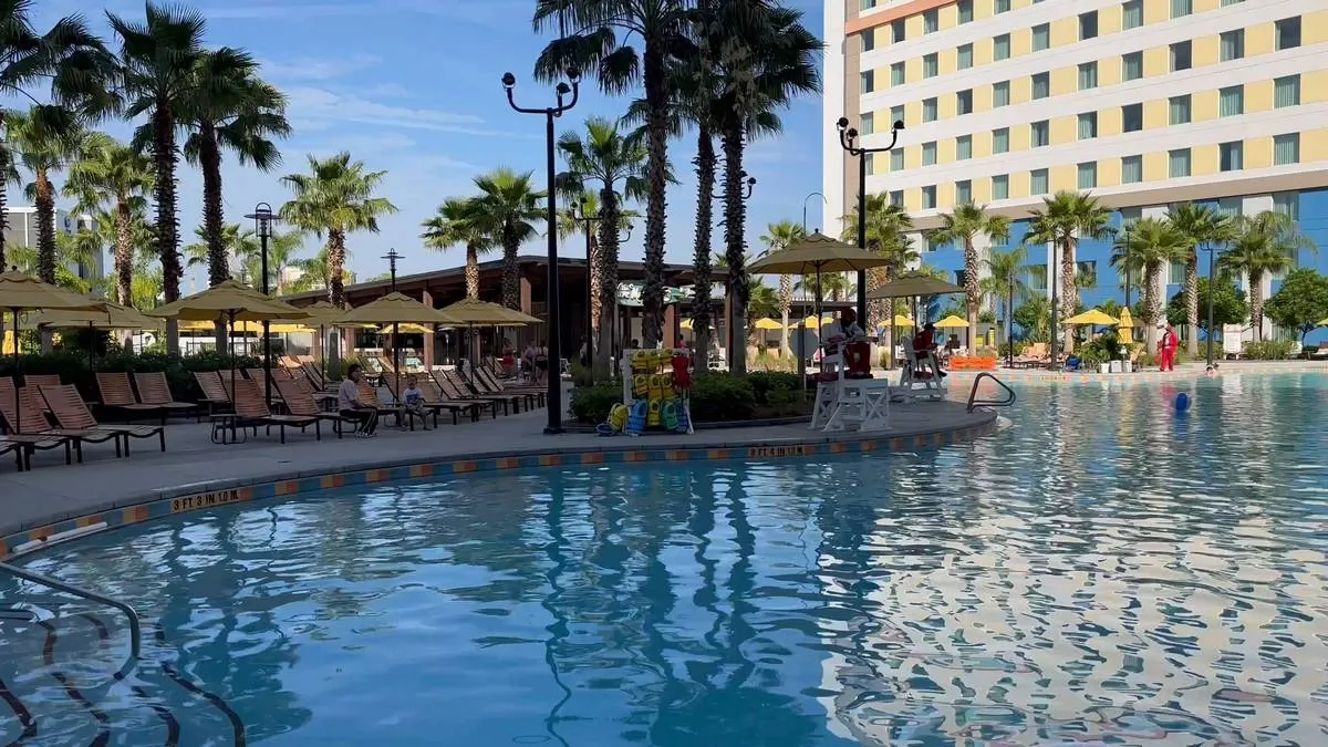 A serene pool area is surrounded by tall palm trees and lined with lounge chairs under yellow umbrellas. A modern hotel building with a colorful facade stands in the background, reflecting in the calm water. The setting is bright and inviting, with a clear blue sky overhead.