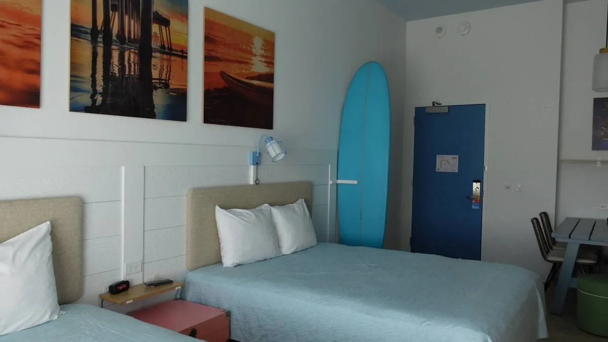 A hotel room features two beds with light blue bedspreads and white pillows. A surfboard leans against the wall near a blue door. A small pink nightstand holds a clock and other items.