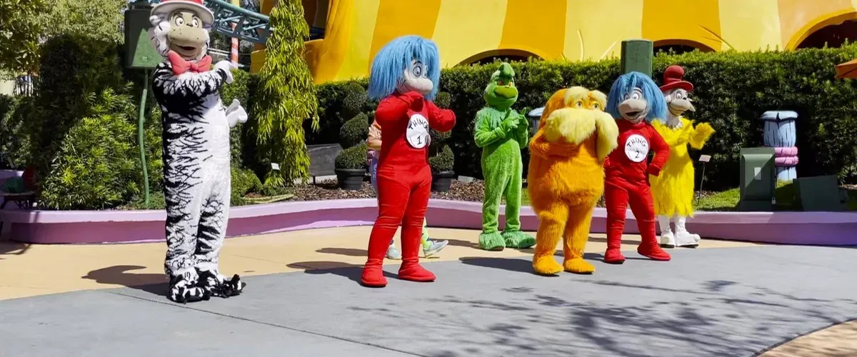 A group of six colorful, costumed characters stands cheerfully on a theme park pathway, surrounded by greenery and whimsical architecture.