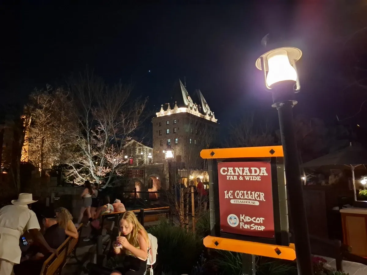 A nighttime scene at a theme park with people sitting on benches and a large illuminated sign reading "Canada Far & Wide," "Le Cellier Steakhouse," and "Kidcot Fun Stop." The background features a lit-up building resembling a castle, surrounded by trees and lamp posts.