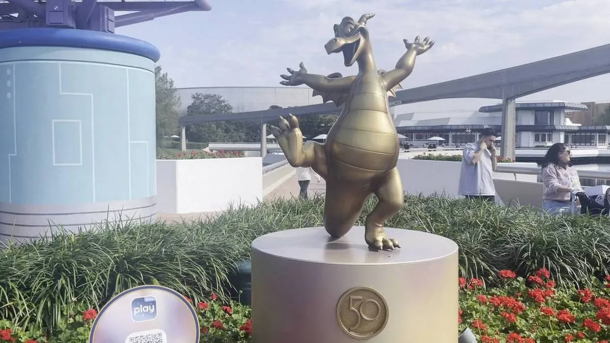 A golden statue of a cheerful dragon stands on a circular pedestal adorned with a "50" emblem, surrounded by greenery and red flowers, with a futuristic blue structure and a monorail track in the background.