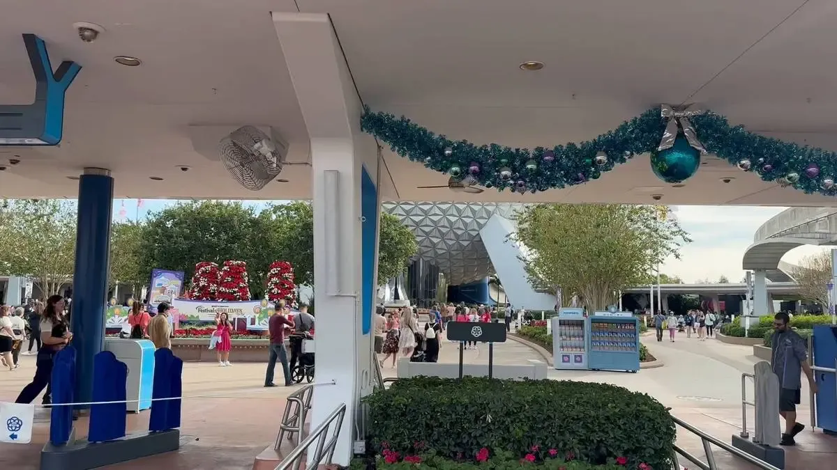 A busy entrance area at a theme park features festive holiday decorations, including a garland with blue ornaments. In the background, a large geodesic sphere is visible, surrounded by trees and visitors walking around.