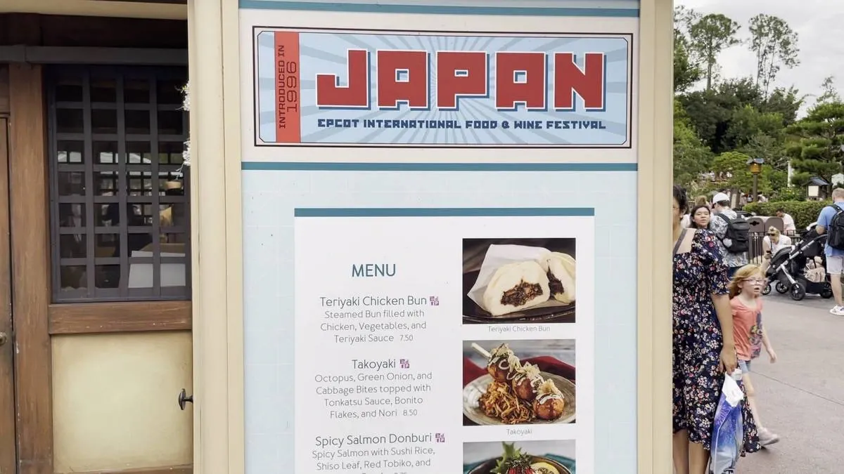 A menu board at the Epcot International Food & Wine Festival in the Japan section features dishes like Teriyaki Chicken Bun and Takoyaki, with images of the food. People walk by in the background, enjoying the outdoor setting.