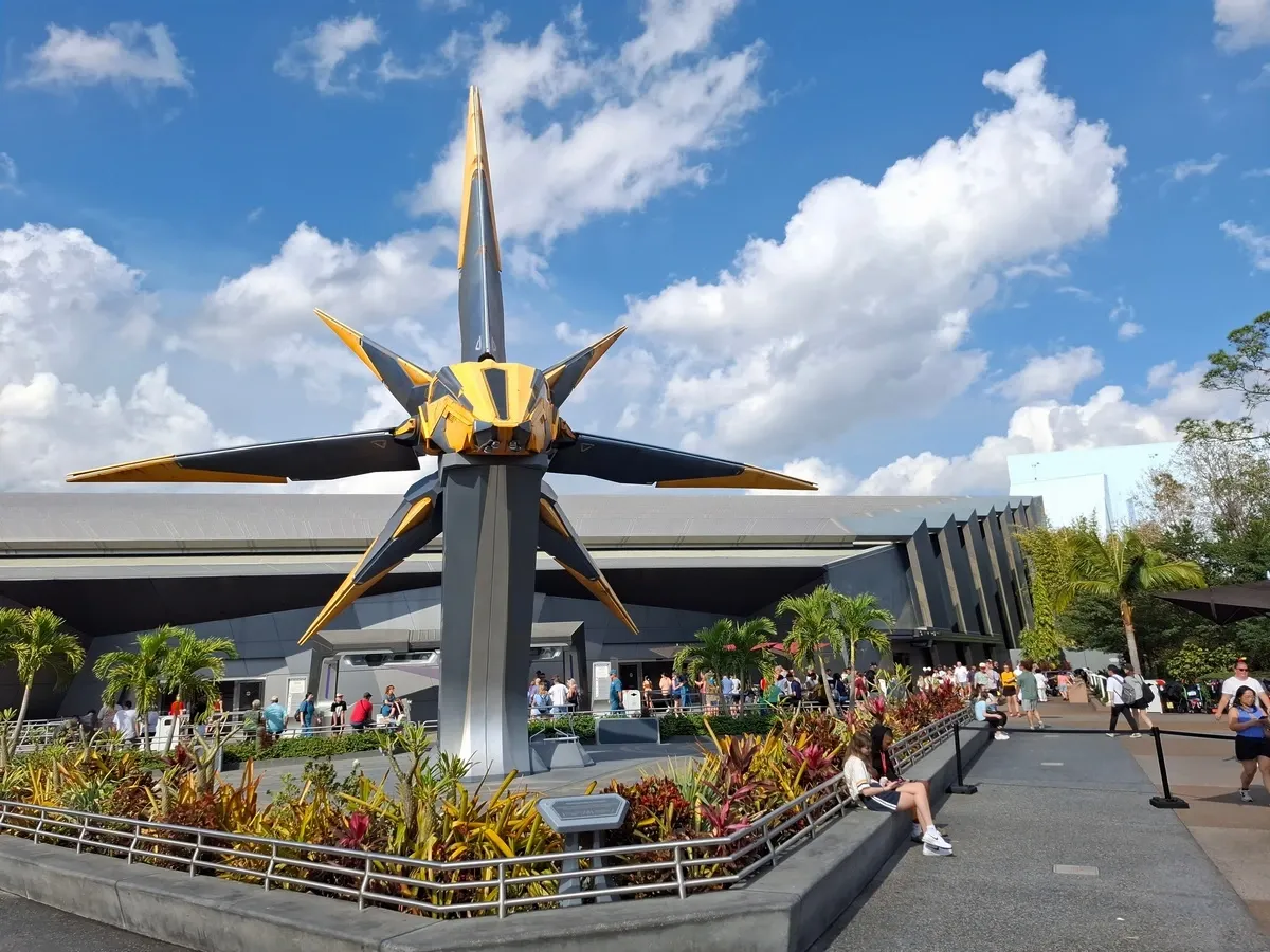 A large, futuristic spacecraft model with sharp, angular wings is prominently displayed outdoors, surrounded by lush plants and a crowd of people. The sky is bright with fluffy clouds, and the building in the background has a sleek, modern design.