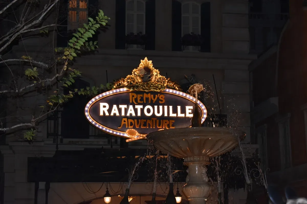 A glowing sign for "Remy's Ratatouille Adventure" is illuminated at night, surrounded by decorative gold accents. In the foreground, a fountain is gently spraying water, adding a lively touch to the scene.