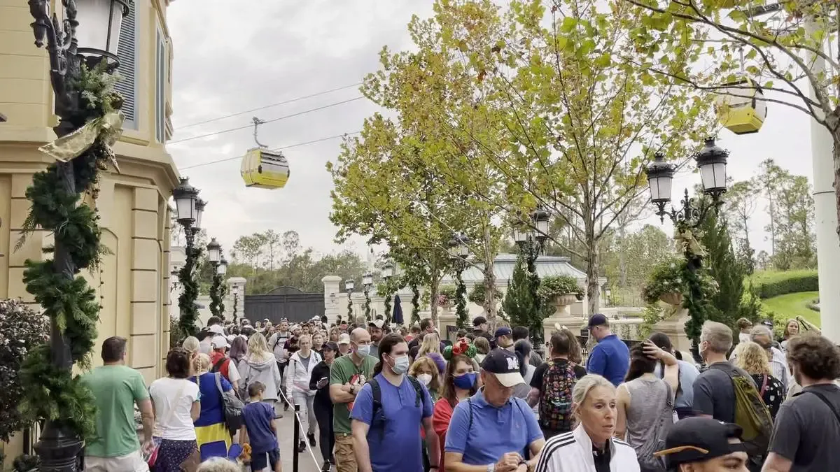 A large crowd of people, some wearing masks, are gathered in an outdoor area with trees and decorative lamp posts. A yellow gondola is visible in the sky, part of a cable car system. The scene has a festive atmosphere with greenery and ribbons adorning the lamp posts.