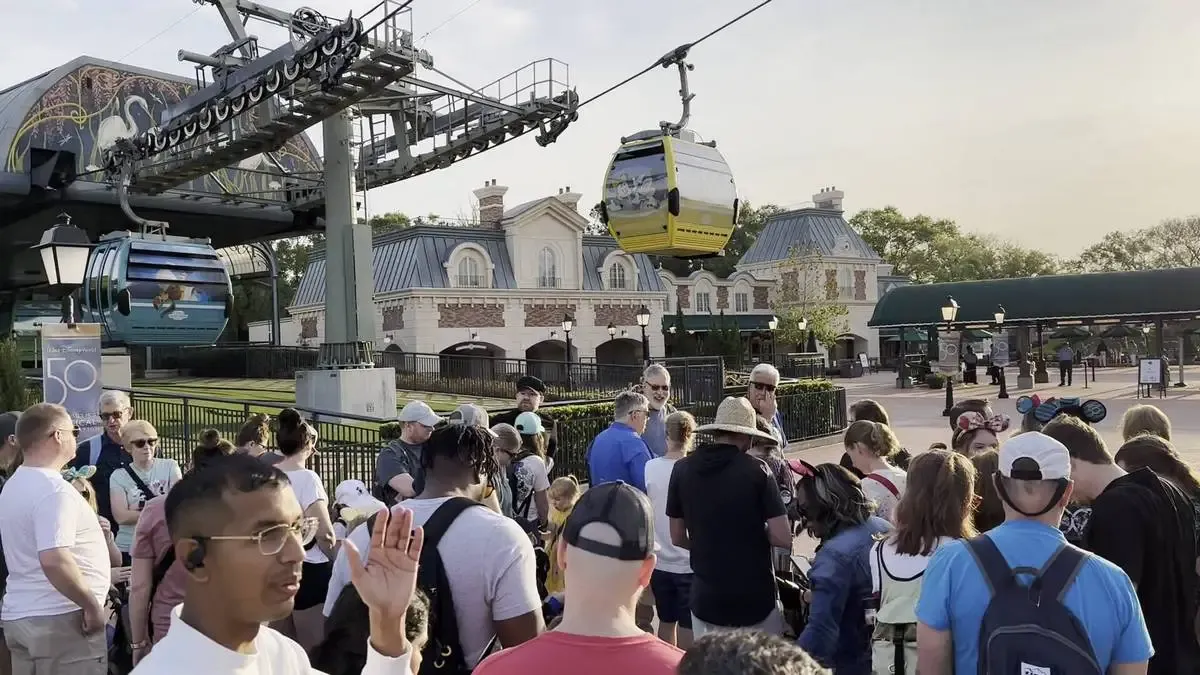 A group of people gathers near a Disney Skyliner gondola station, with colorful gondolas suspended overhead. The background features a building with a classic architectural style, and the scene is set in a sunny, outdoor area.