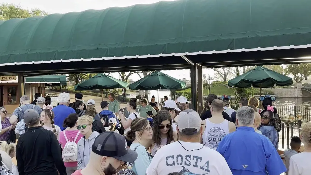 A large crowd of people gathers under a green awning, some wearing backpacks and hats, with a few children visible. Green umbrellas are seen in the background, and the scene appears lively and bustling.