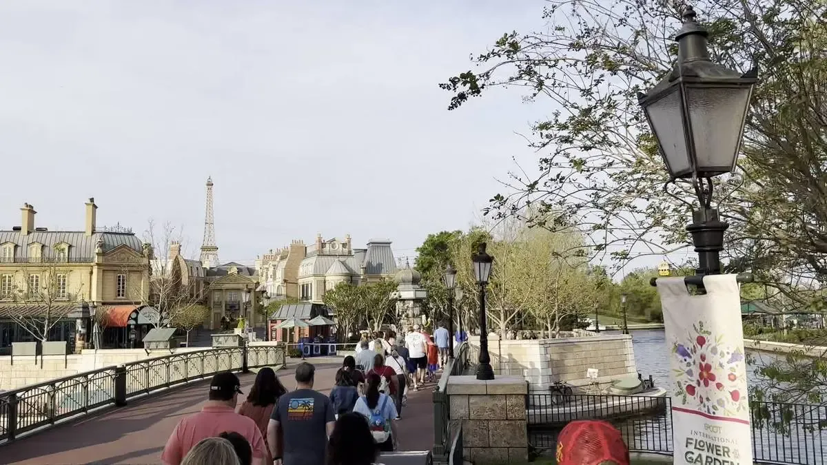 A group of people walk along a path lined with street lamps, with buildings resembling Parisian architecture in the background. A replica of the Eiffel Tower is visible, and a banner on the right reads "EPCOT International Flower & Garden Festival.".