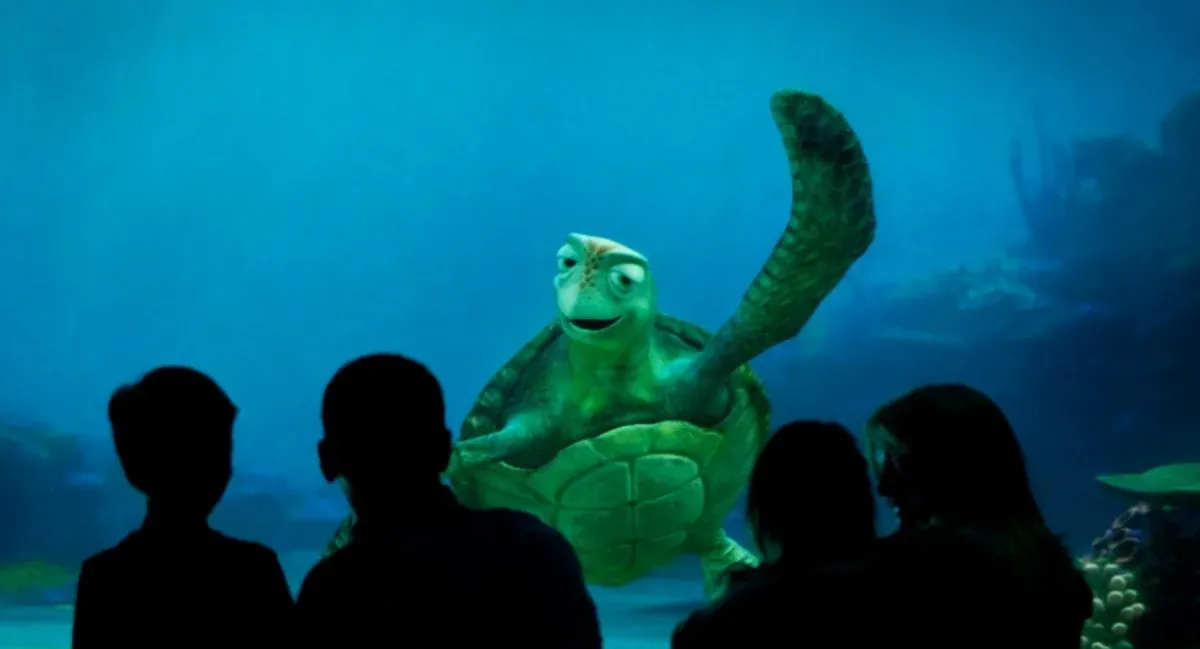 A group of silhouetted people watch an animated green sea turtle with a friendly expression swimming in a large aquarium, set against a deep blue underwater background.