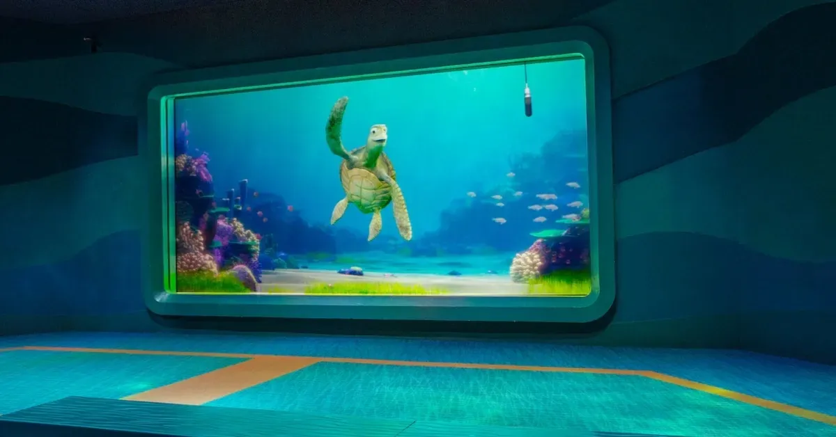 An animated sea turtle swims in a large aquarium display, surrounded by colorful coral and a school of fish, with a microphone hanging above the tank.