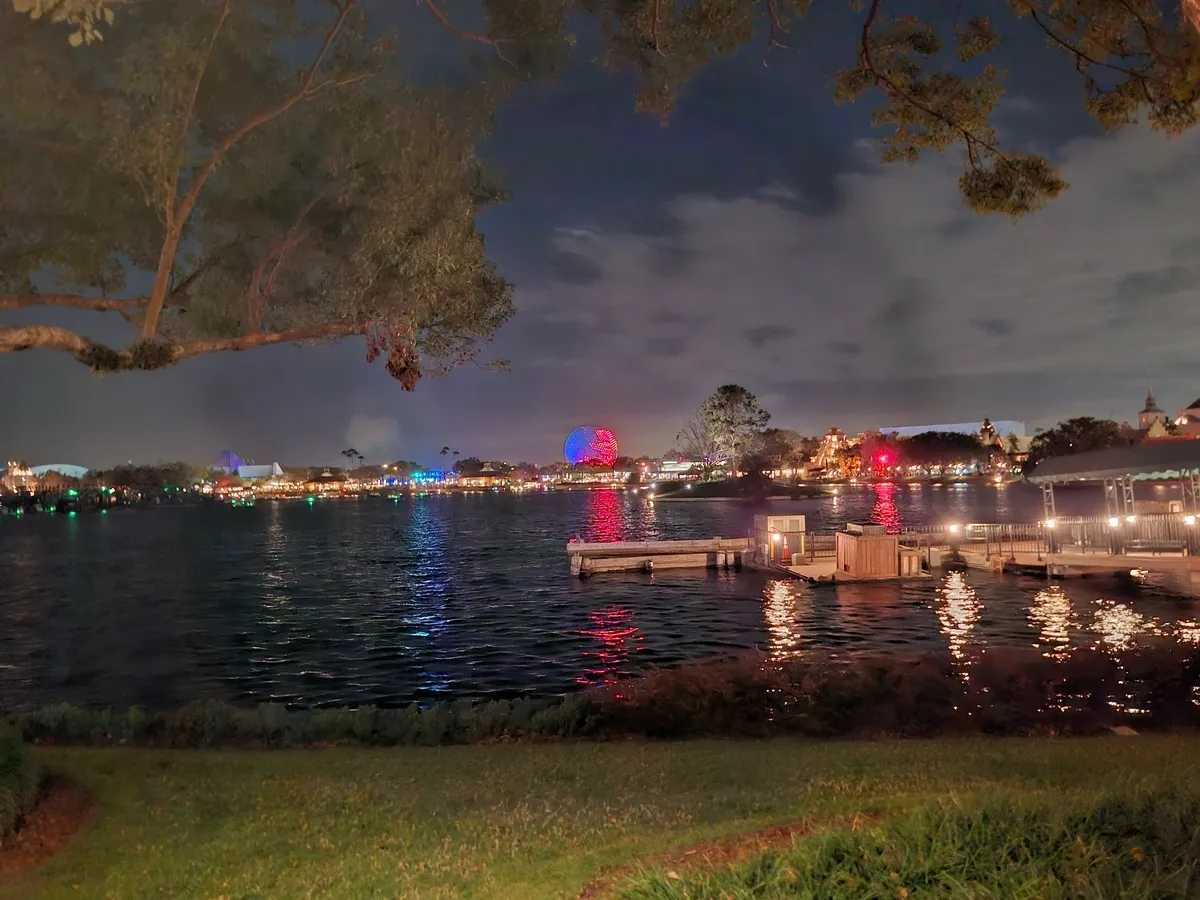A serene nighttime scene shows a waterfront with shimmering reflections of colorful lights on the water. In the background, a large, illuminated sphere glows in red and blue hues, surrounded by distant buildings.