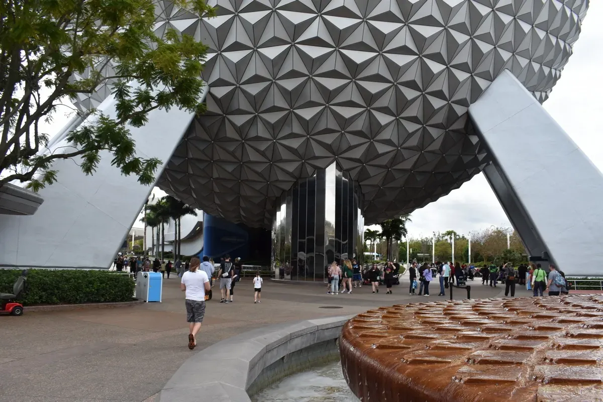 People walk around the base of the iconic geodesic sphere at Epcot, with a fountain in the foreground and trees nearby, creating a lively and bustling atmosphere.