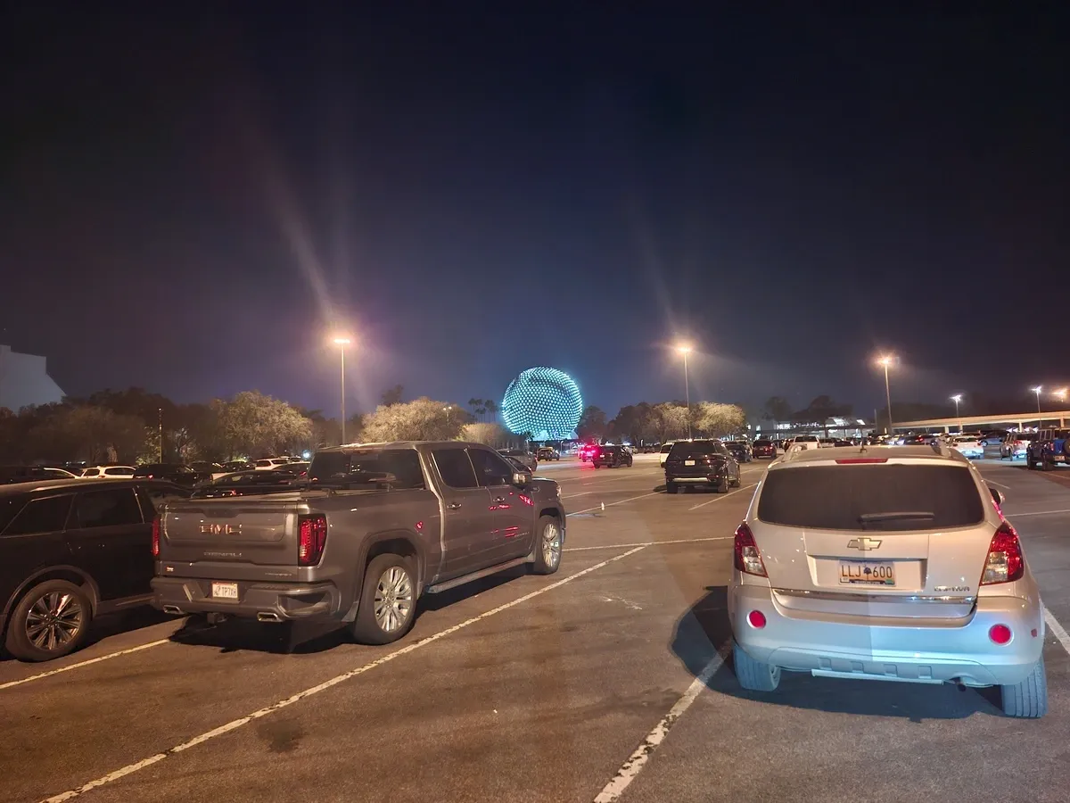 A parking lot at night with various vehicles, including a GMC truck and a Chevrolet SUV, under streetlights. In the background, a large illuminated geodesic sphere is visible, surrounded by trees.