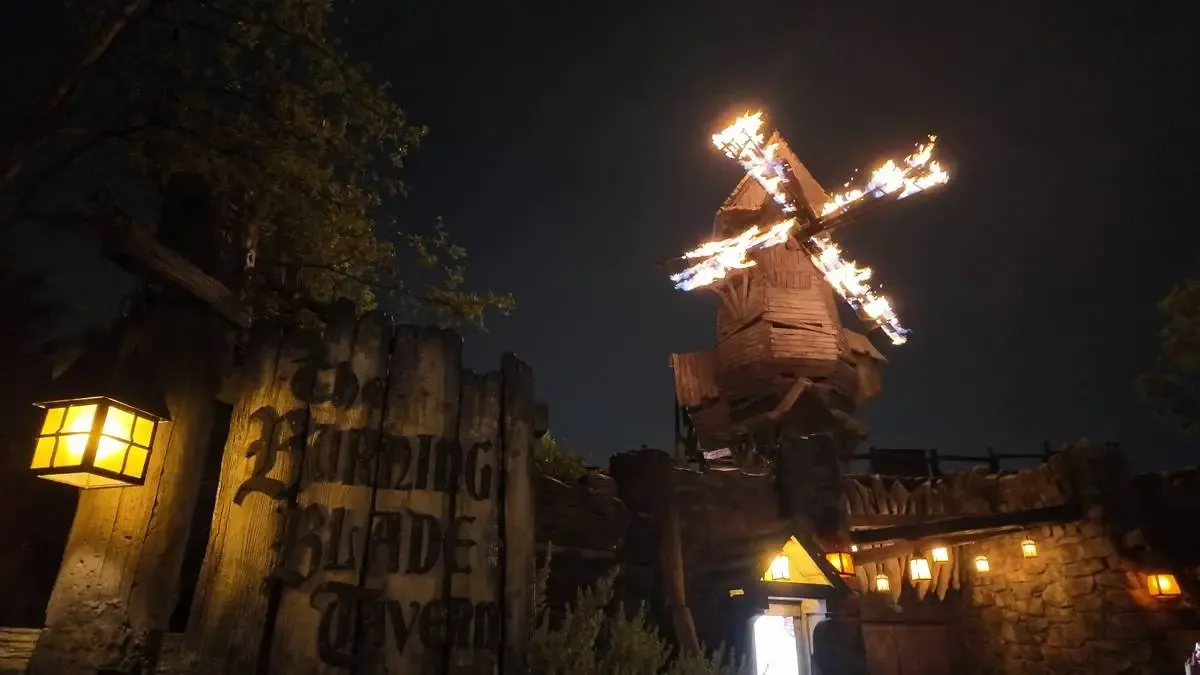 A wooden windmill with flaming blades illuminates the night sky above a rustic tavern sign reading "The Burning Blade Tavern," surrounded by glowing lanterns and trees.