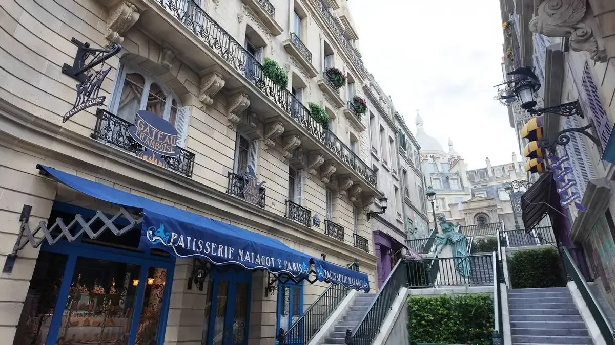 A charming Parisian street scene features a patisserie with a blue awning displaying "Patisserie Matagot y Paris." The elegant building has ornate balconies with flower boxes, and a staircase with greenery leads up to a statue.