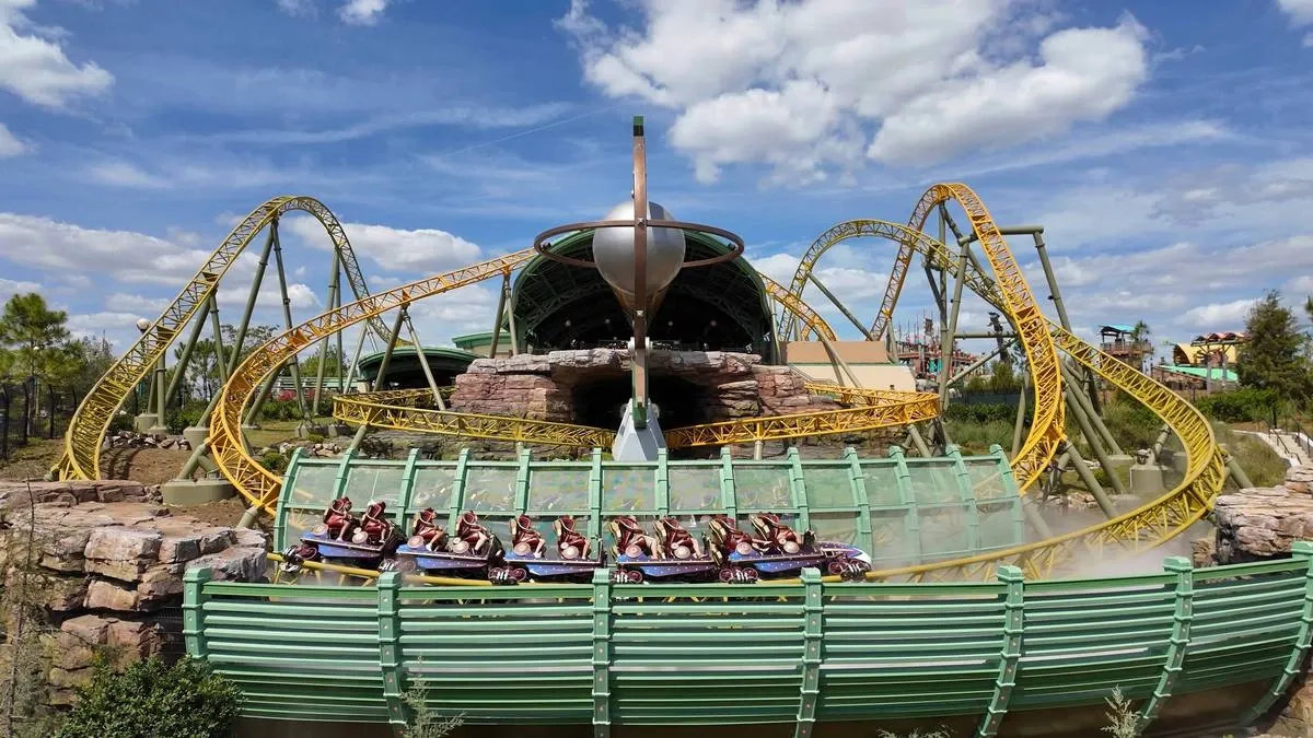 A vibrant roller coaster with a yellow track twists and loops around a rocky landscape, with a train full of excited riders in motion. The setting is under a bright blue sky with scattered clouds, adding to the lively amusement park atmosphere.