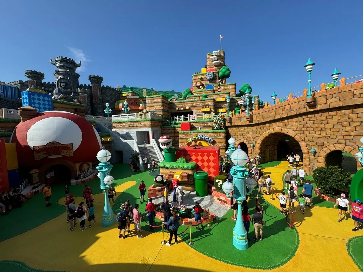 Visitors explore a theme park area inspired by a Mario Bros video game, featuring colorful structures like a giant mushroom and a castle. The scene is bustling with people walking along bright yellow and green paths under a clear blue sky.