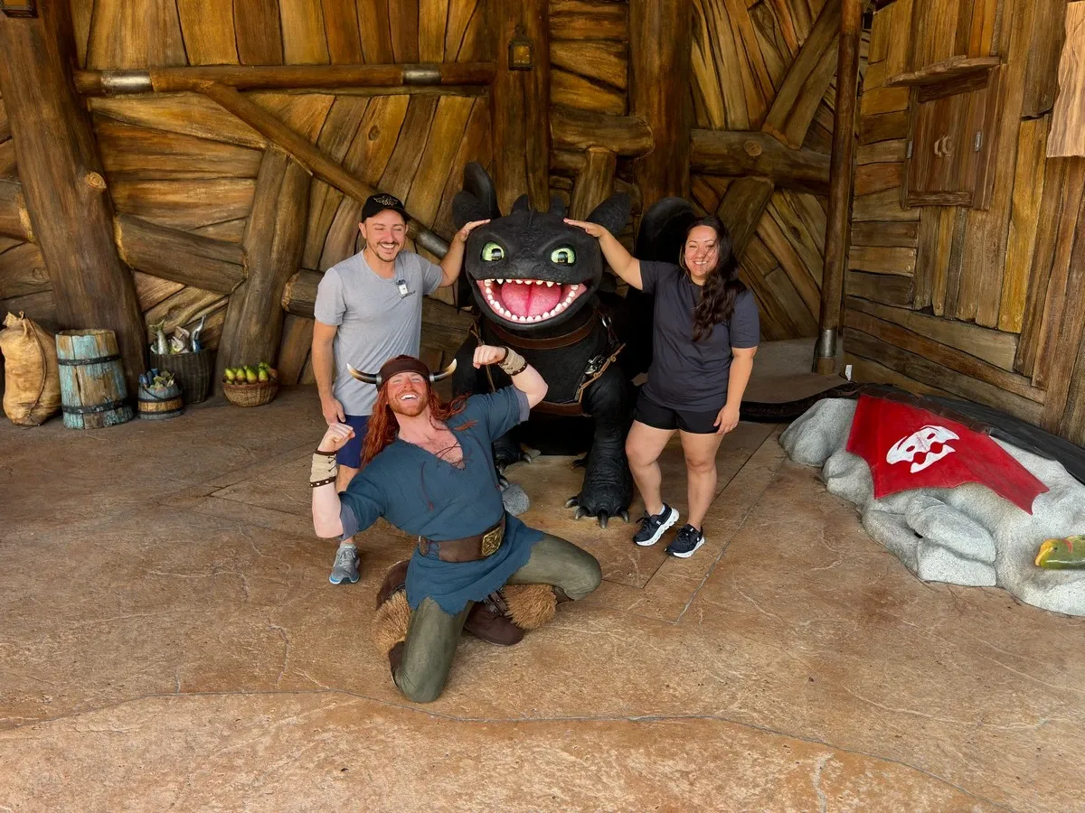 Two people pose with a large dragon figure inside a rustic wooden setting, while a costumed character kneels playfully in front, flexing his arms. A red flag with a white design is visible on the ground.
