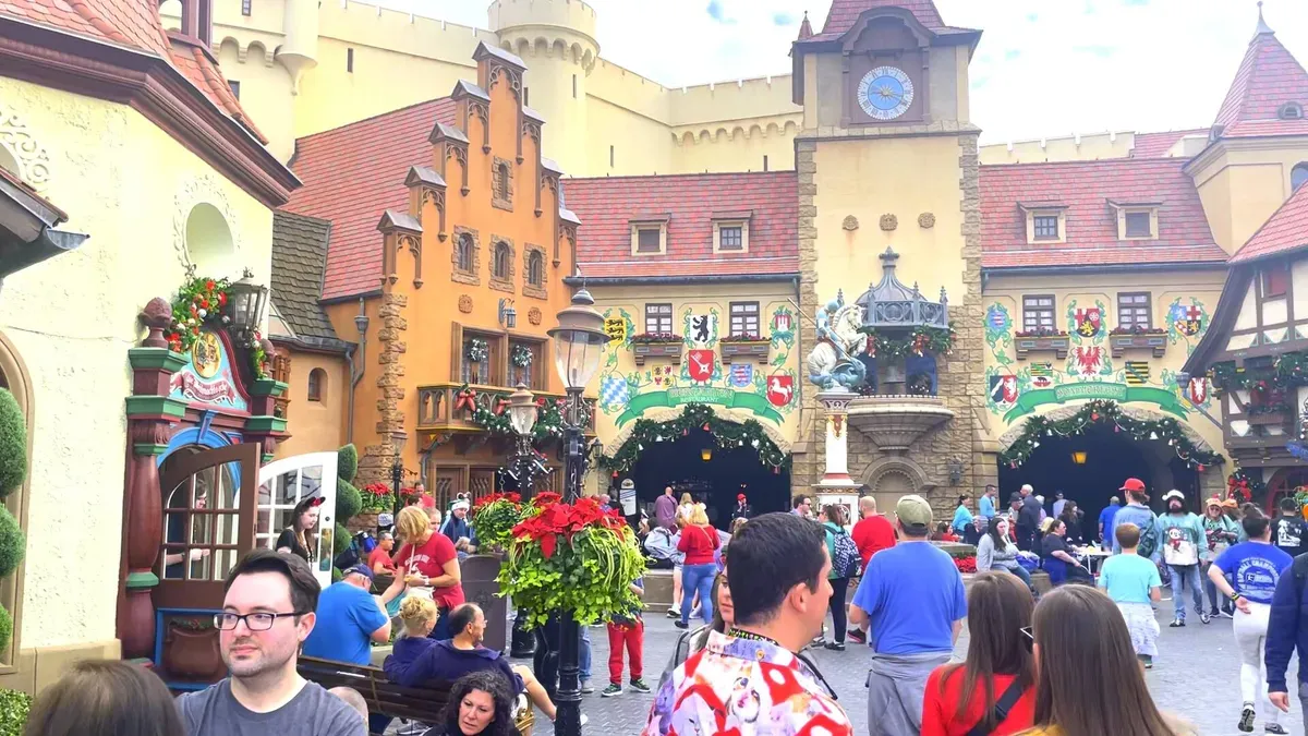 Scene at a theme park showcases an area with colorful, German-style architecture. Crowds of people, some in festive attire, gather around a central fountain adorned with statues. 