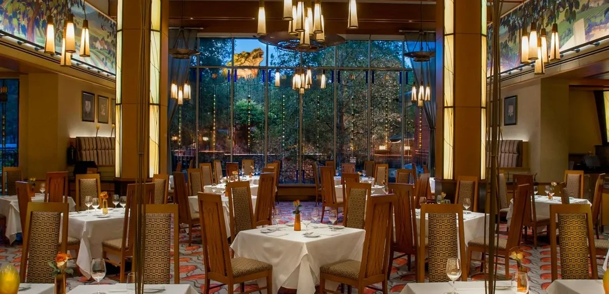 A warm restaurant interior featuring tables with white tablecloths and wooden chairs. Large windows offer a view of greenery outside, while modern pendant lights hang from the ceiling.