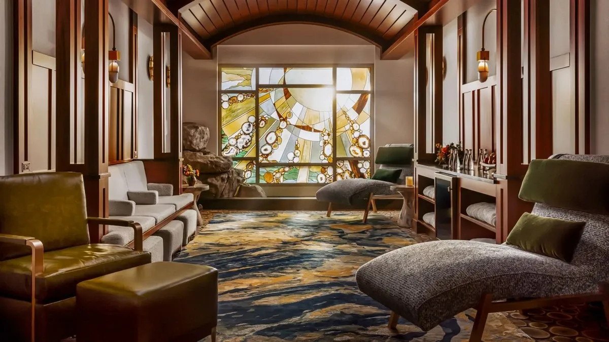 A spa lounge features plush loungers and armchairs in a warm, inviting setting. A stunning stained glass window with an abstract design allows soft light to filter in, enhancing the elegant atmosphere. The room is adorned with wooden accents, soft lighting, and a colorful patterned carpet.