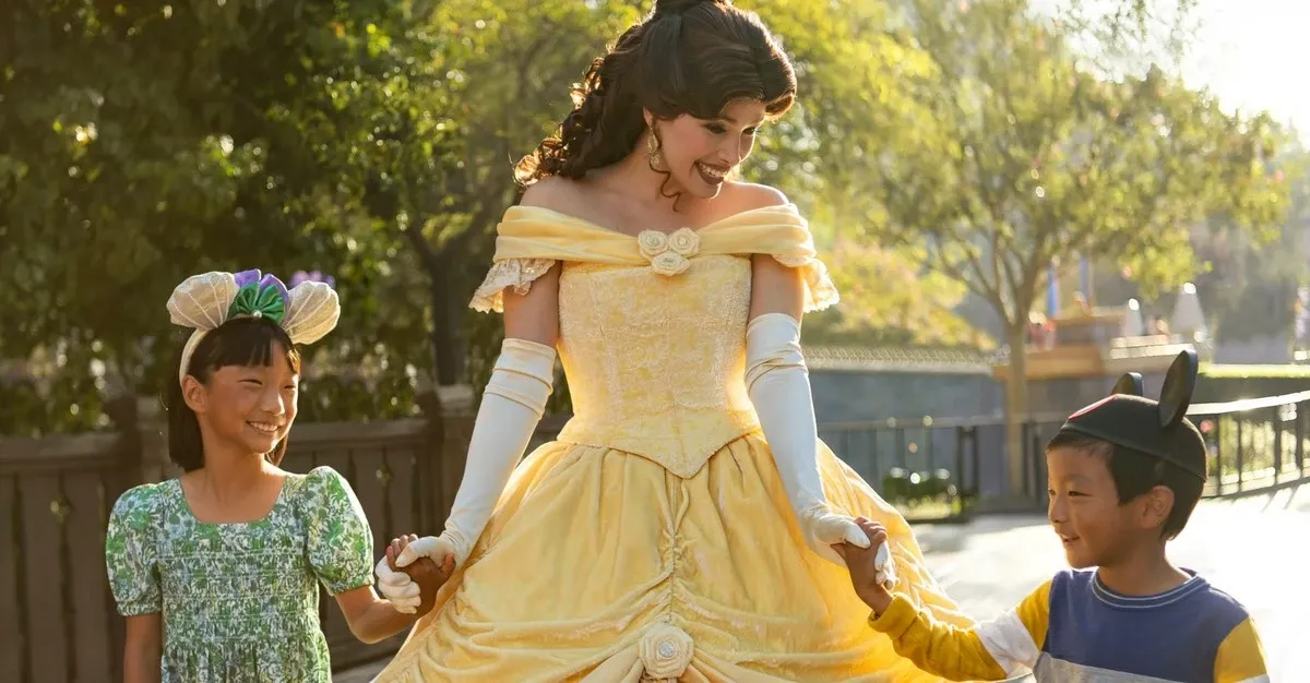 A woman dressed as a Belle from "Beauty and the Beast", joyfully holds hands with two smiling children, one wearing a green dress and floral headband, and the other in a blue shirt with mouse ears.