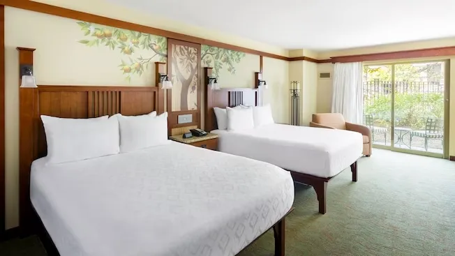A hotel room featuring two double beds with white linens, wooden headboards, and a nature-themed mural on the wall. A cozy armchair sits near a large sliding glass door that opens to a balcony with a view of greenery outside.