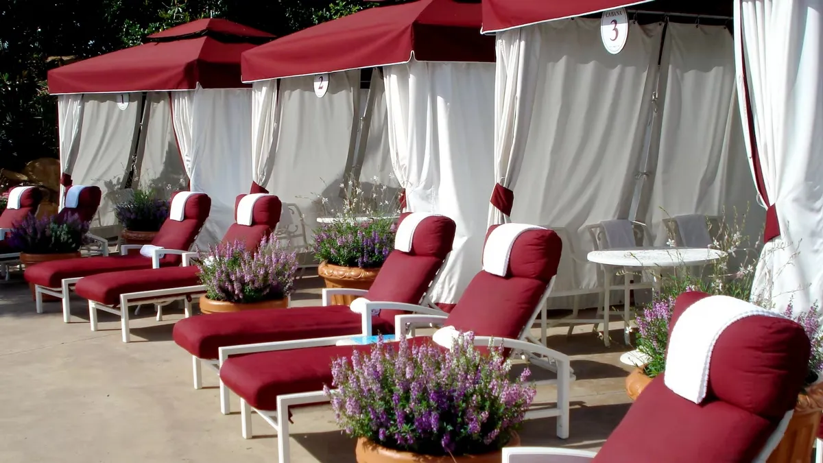 A row of luxurious poolside cabanas with red and white lounge chairs and matching canopies. Each chair is adorned with a white towel and surrounded by potted lavender plants.